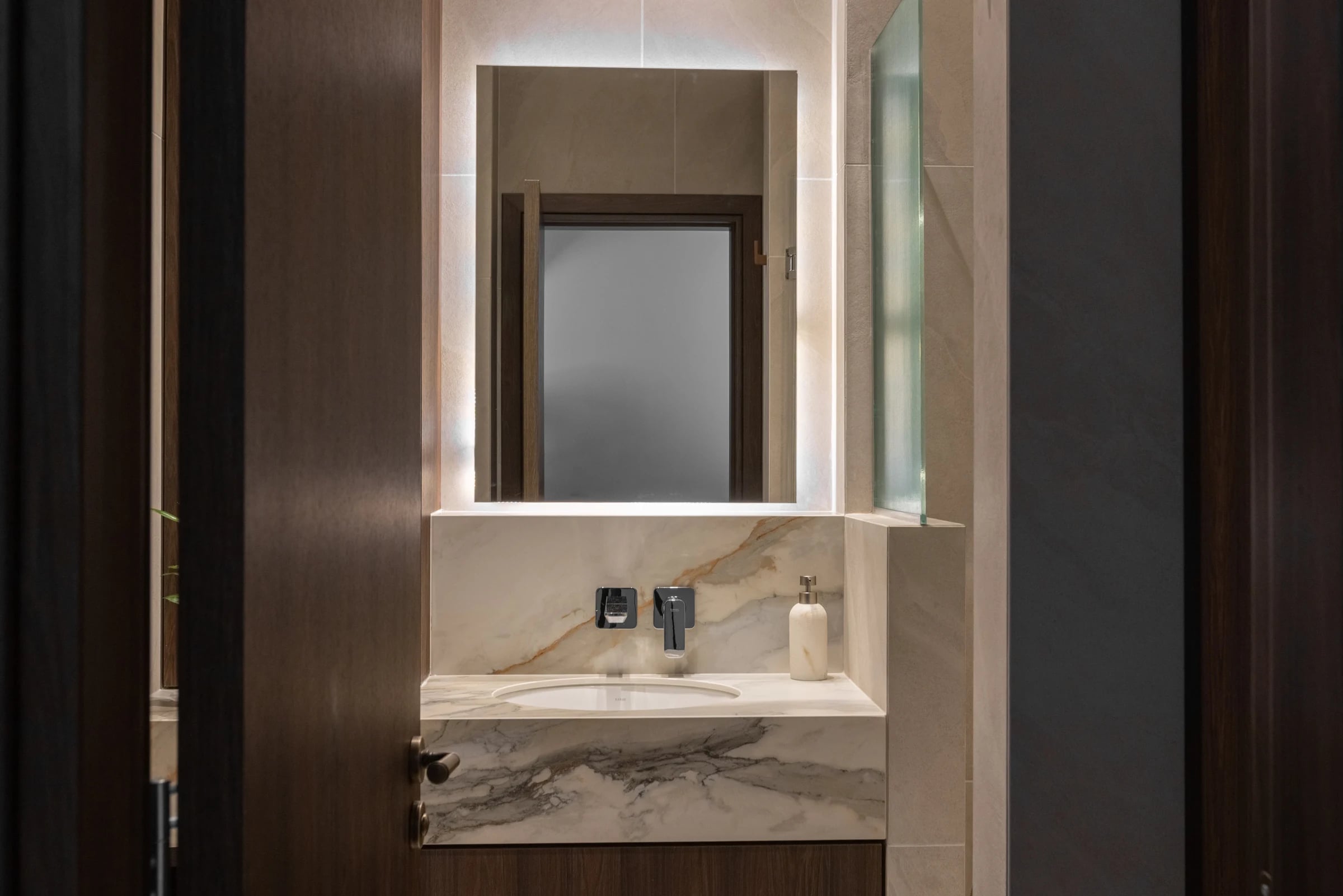 A bathroom vanity with a veined countertop, rectangular illuminated mirror and dark timber cabinetry.