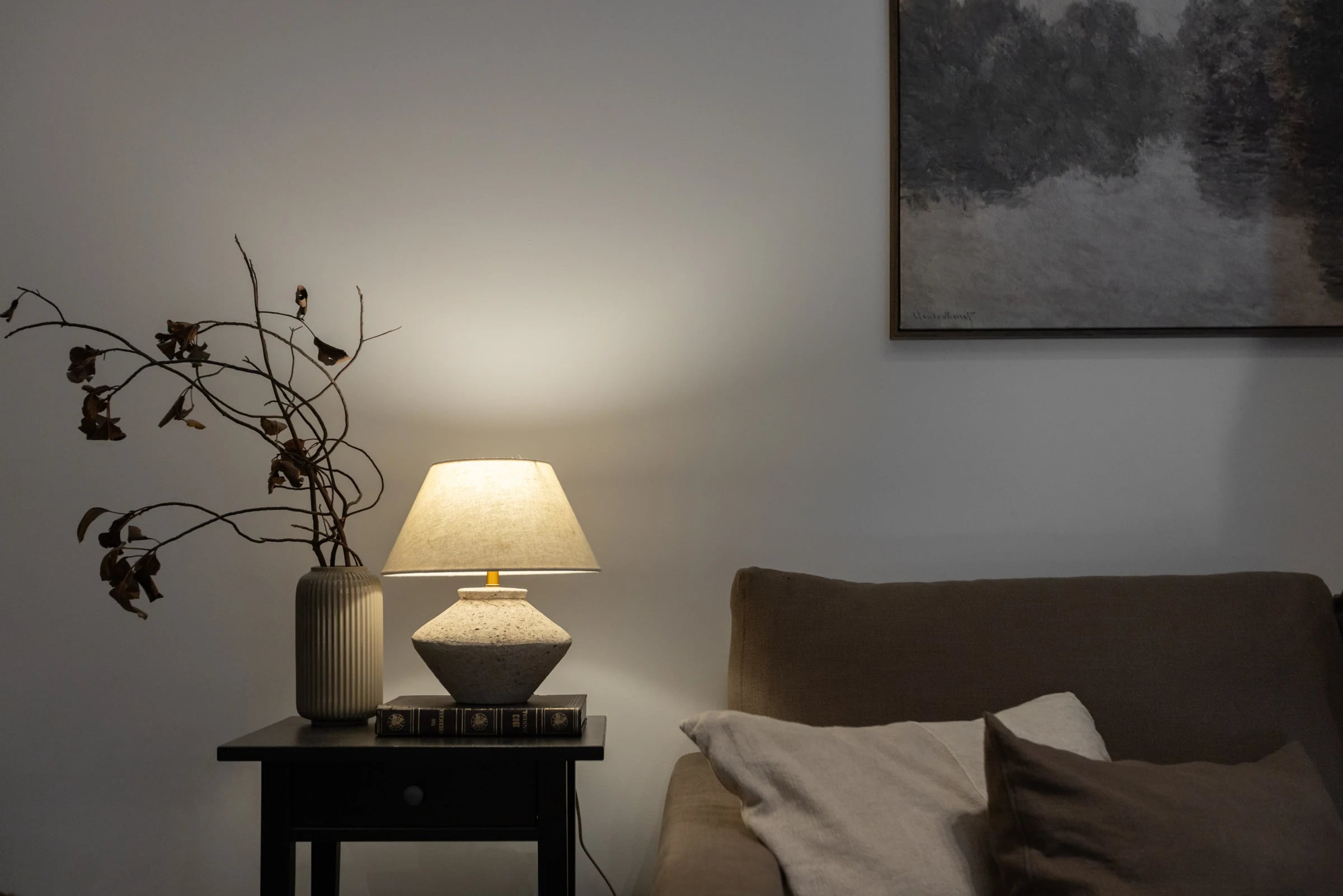 A lit ceramic table lamp and branch-filled vase beside a taupe sofa and framed artwork.