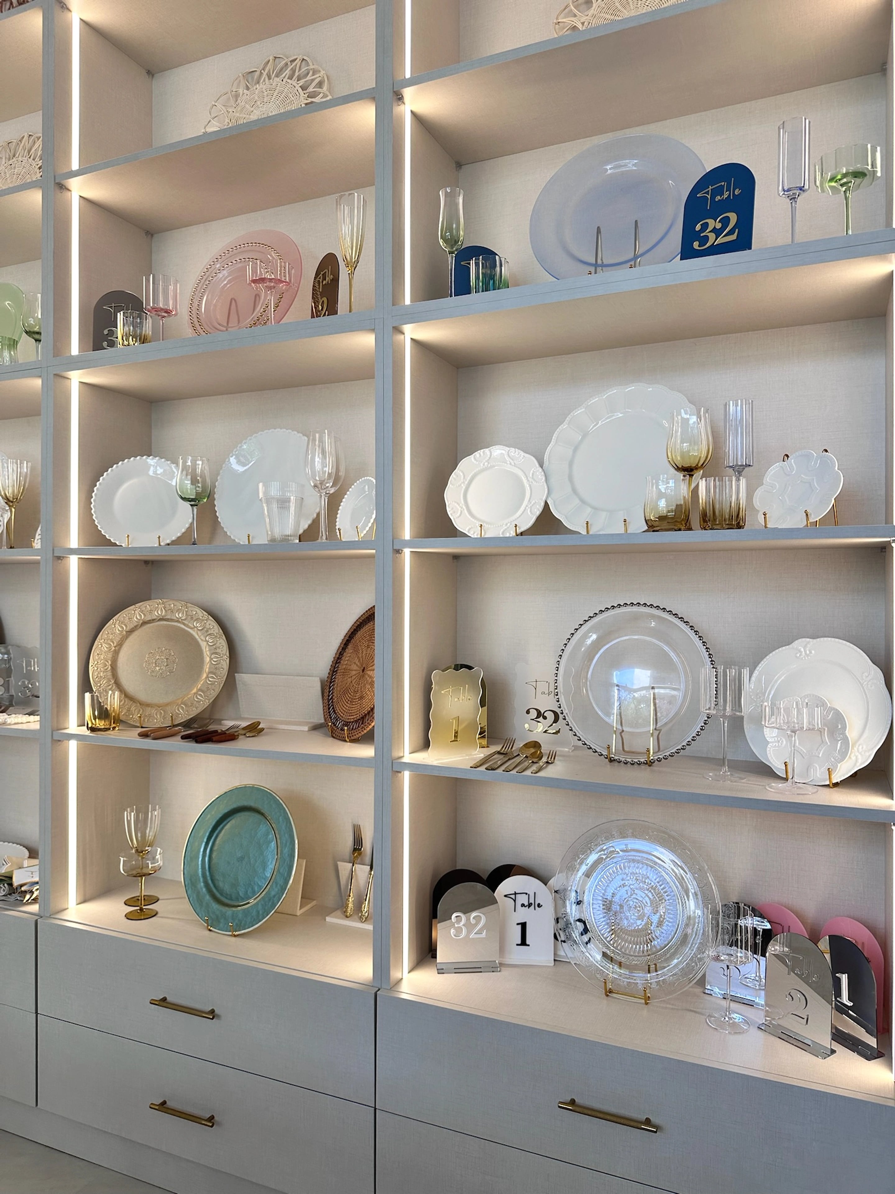 A tall grid of illuminated shelves displaying plates and glassware above fitted drawers.