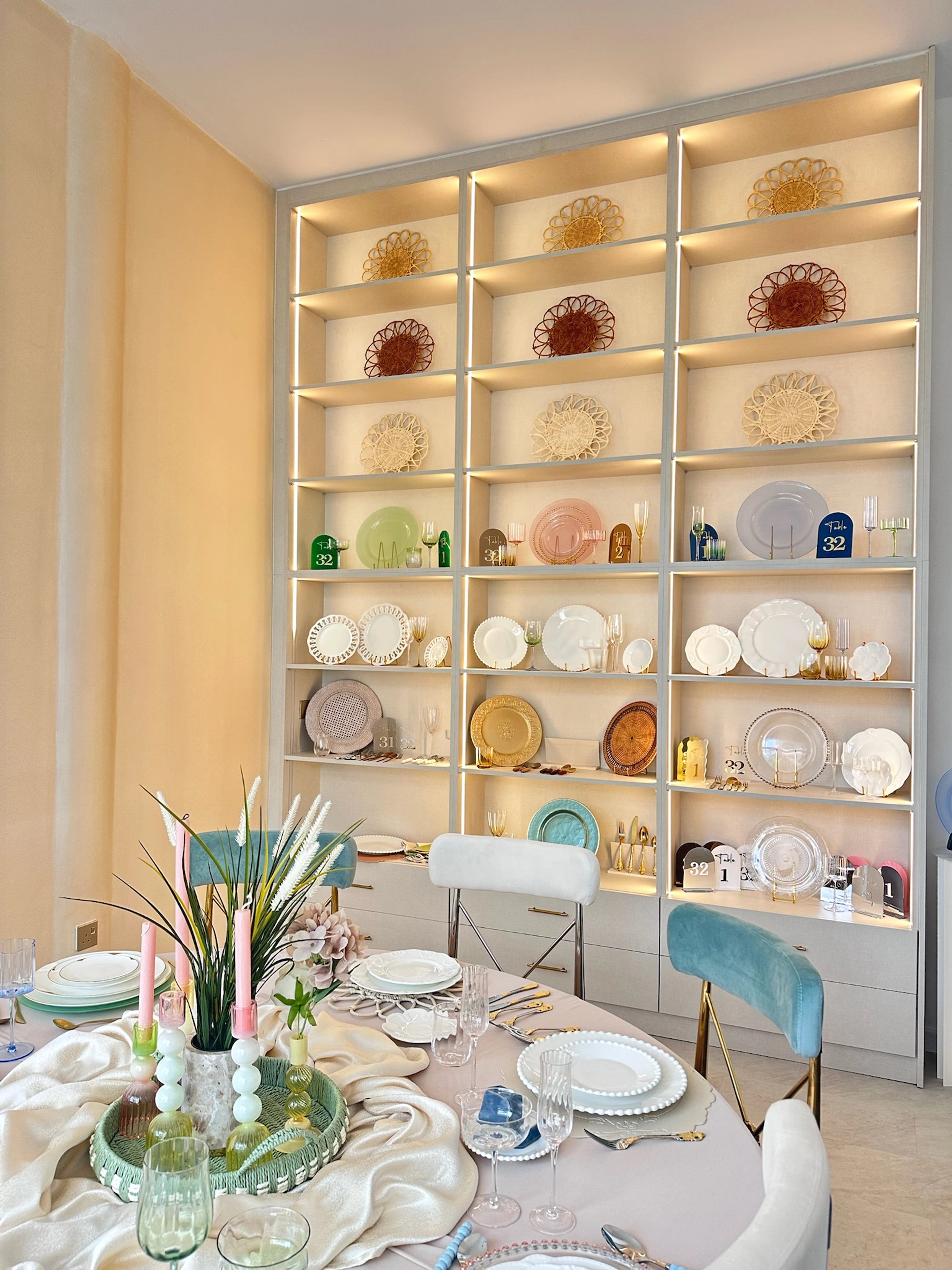 A set dining table with coloured candles in front of illuminated tableware shelves.