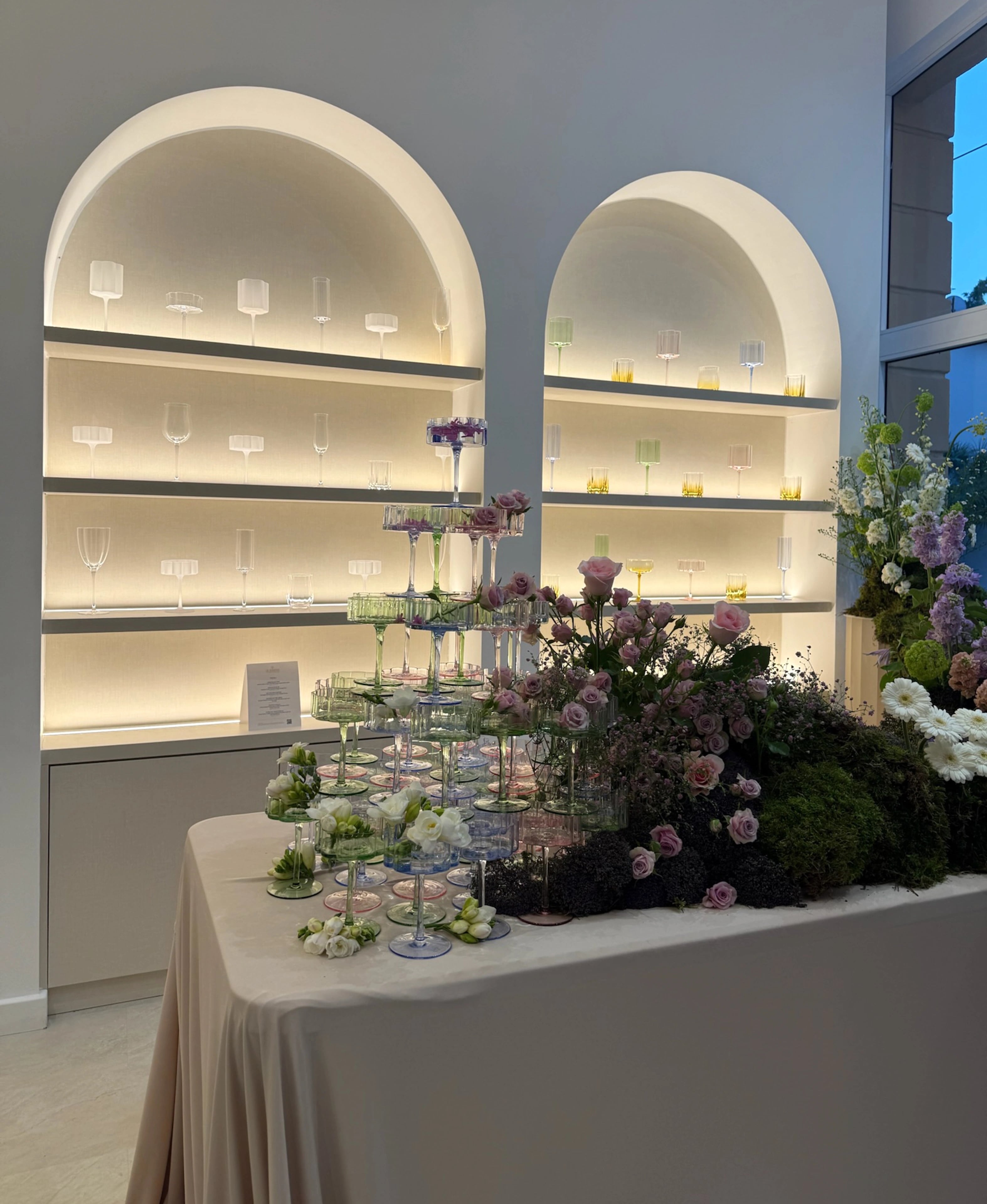 An arranged dining table with flowers in front of illuminated arched display shelves.
