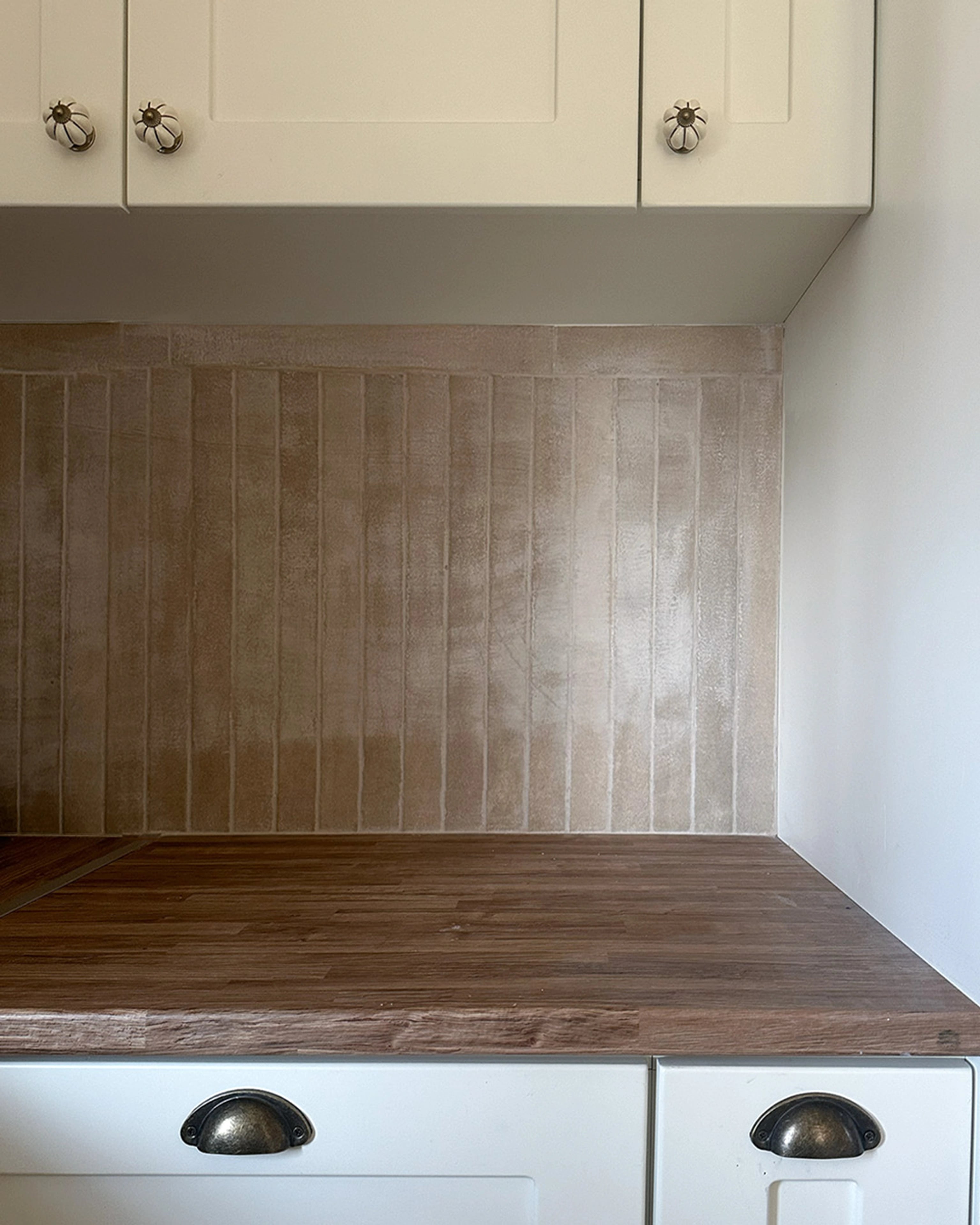 Cream kitchen drawers with dark cup handles beneath a timber worktop and tiled wall.