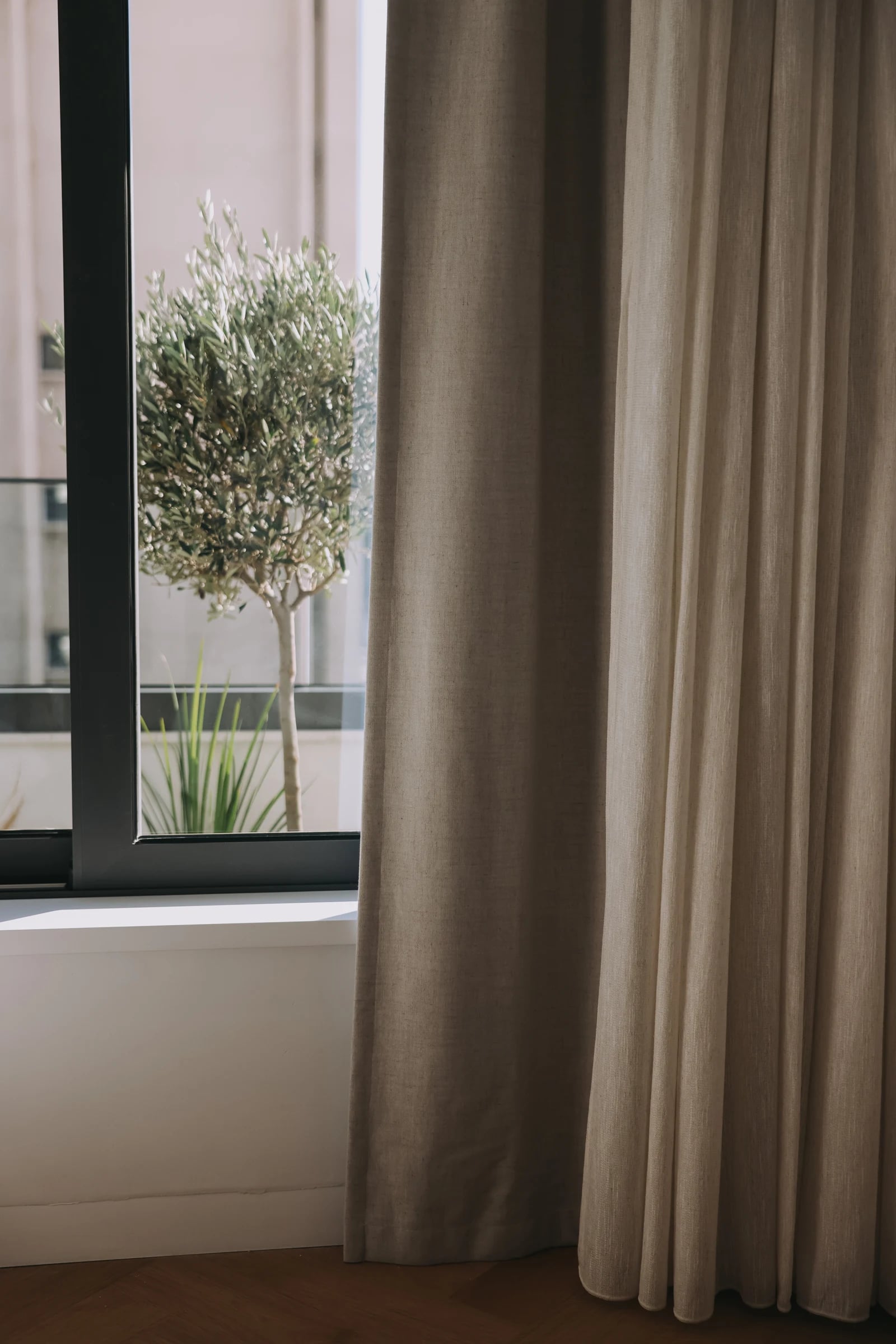 Full-length neutral curtains beside a window overlooking a potted tree.