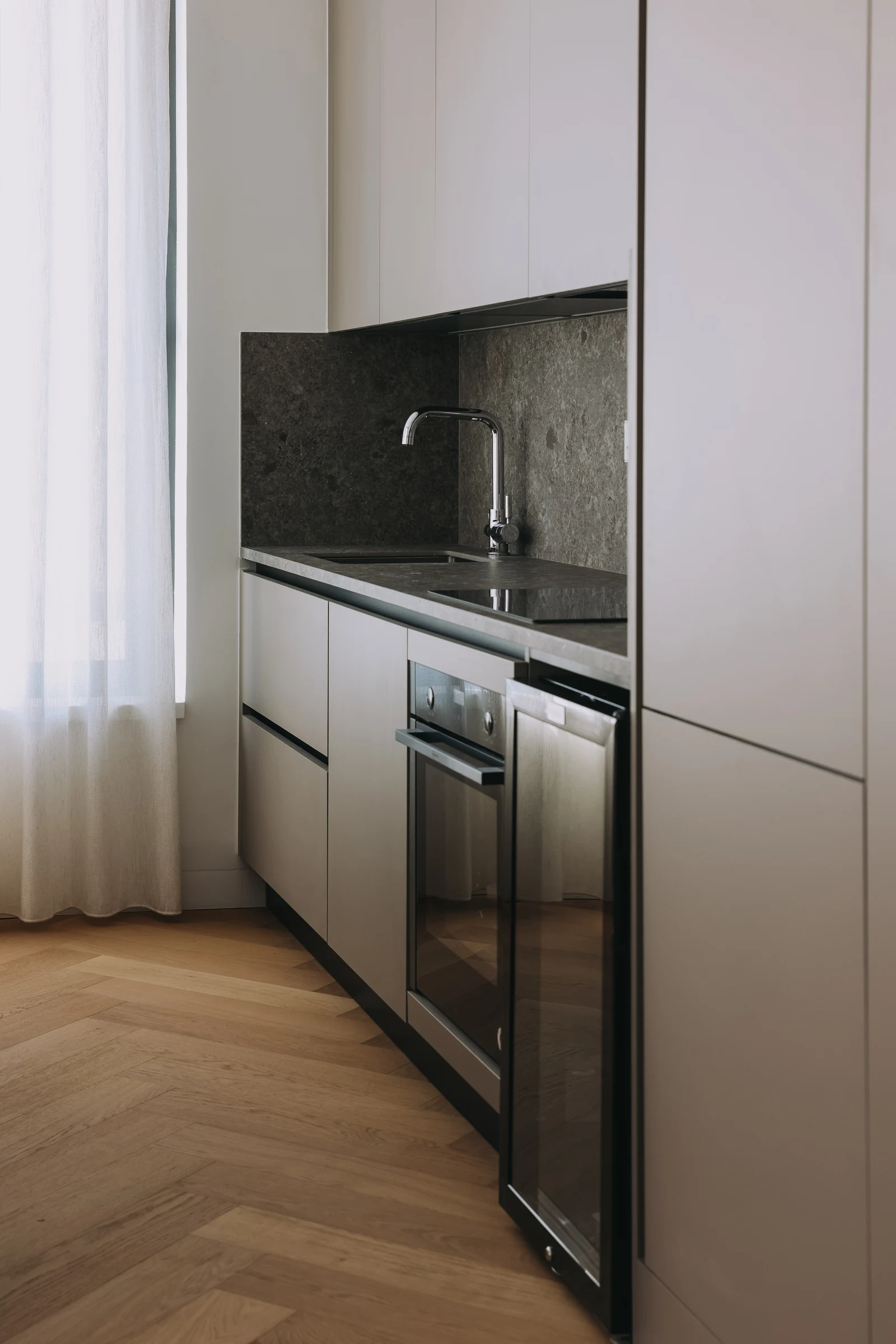 A compact kitchen with pale cabinetry, a dark splashback and herringbone flooring beside a curtain.