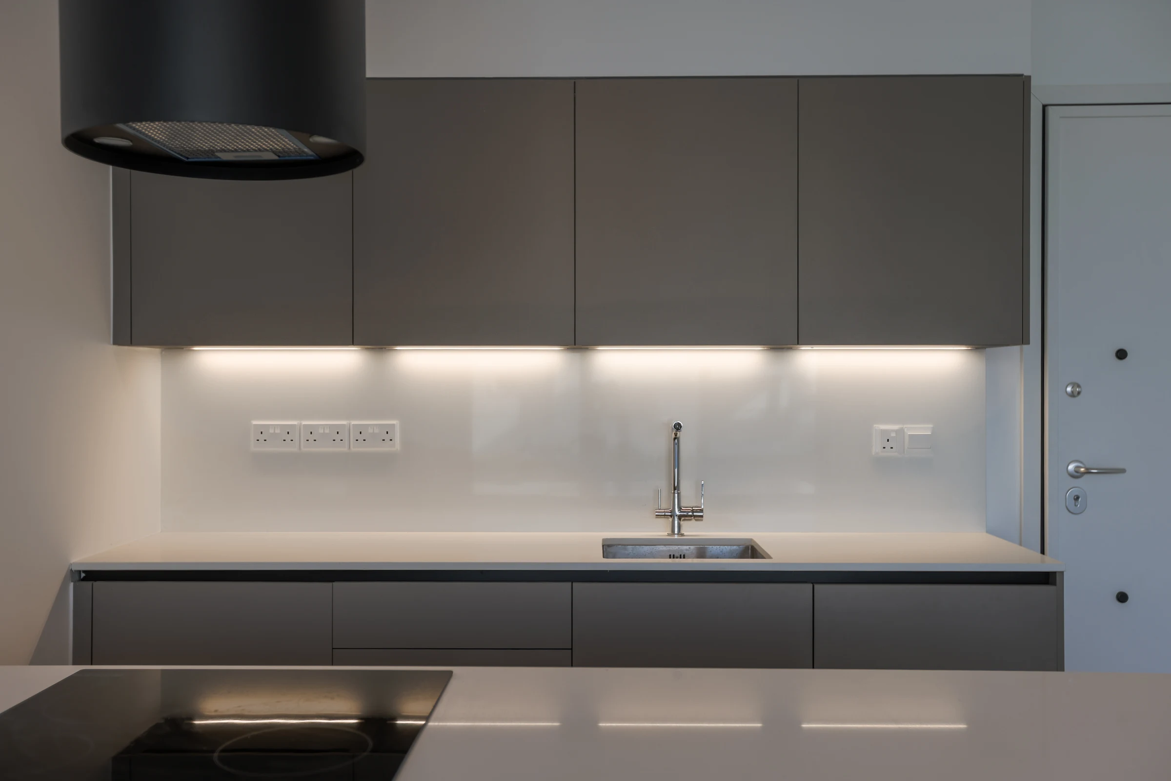 Grey kitchen cabinets and an illuminated white splashback behind a pale island.