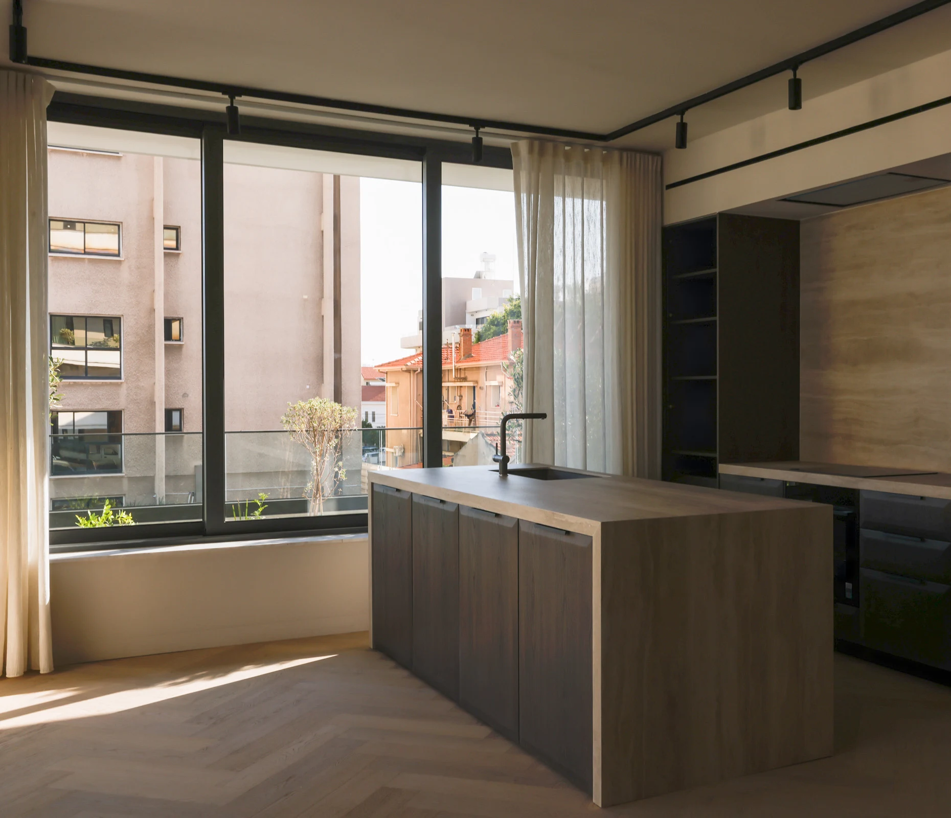Sunlight falling across a timber kitchen island, pale herringbone floor and full-height windows.
