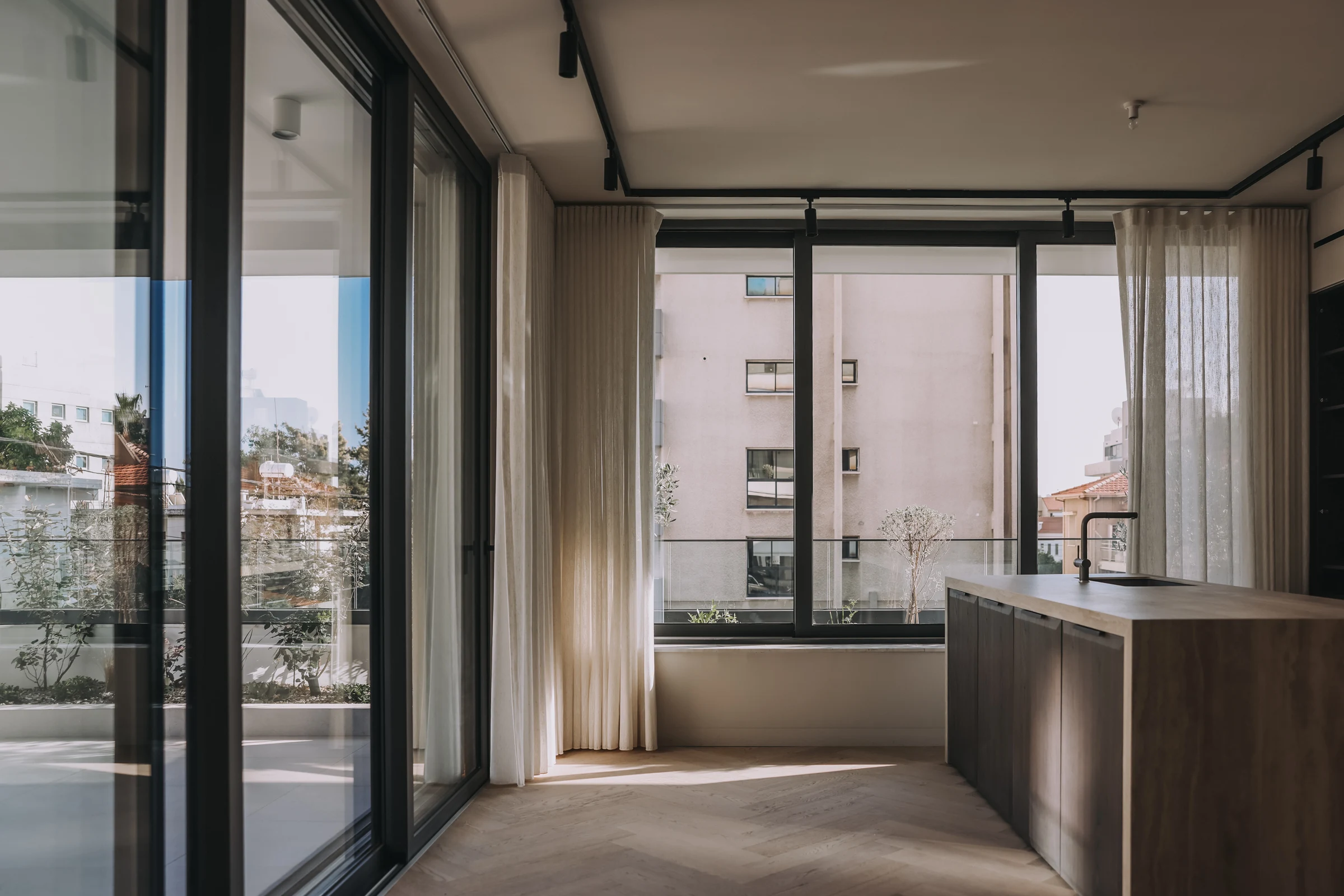 Sunlight entering an unfurnished kitchen through full-height windows beside a timber island.