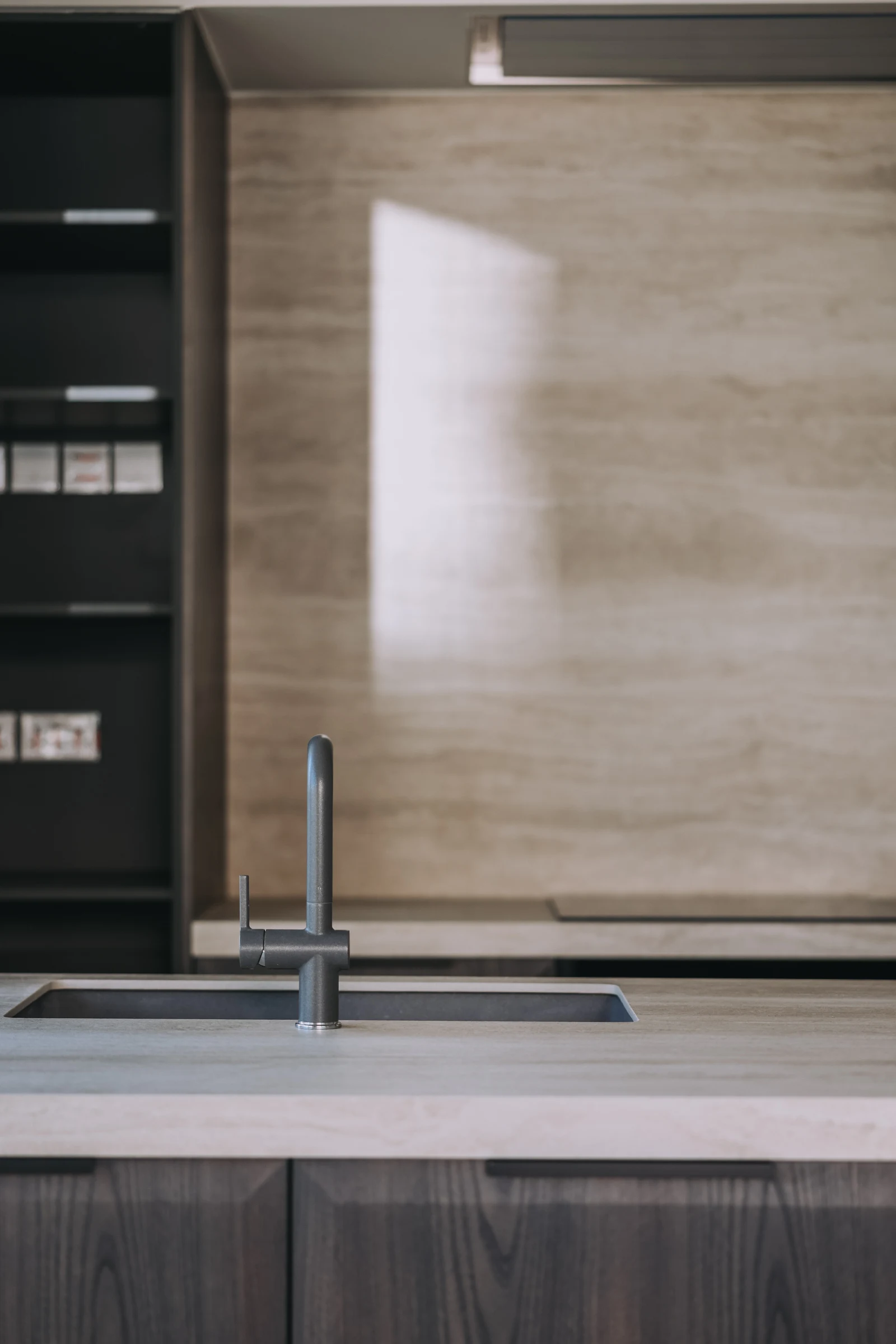 A dark kitchen tap and sink beneath a pale stone-patterned wall panel and slim pendant lights.