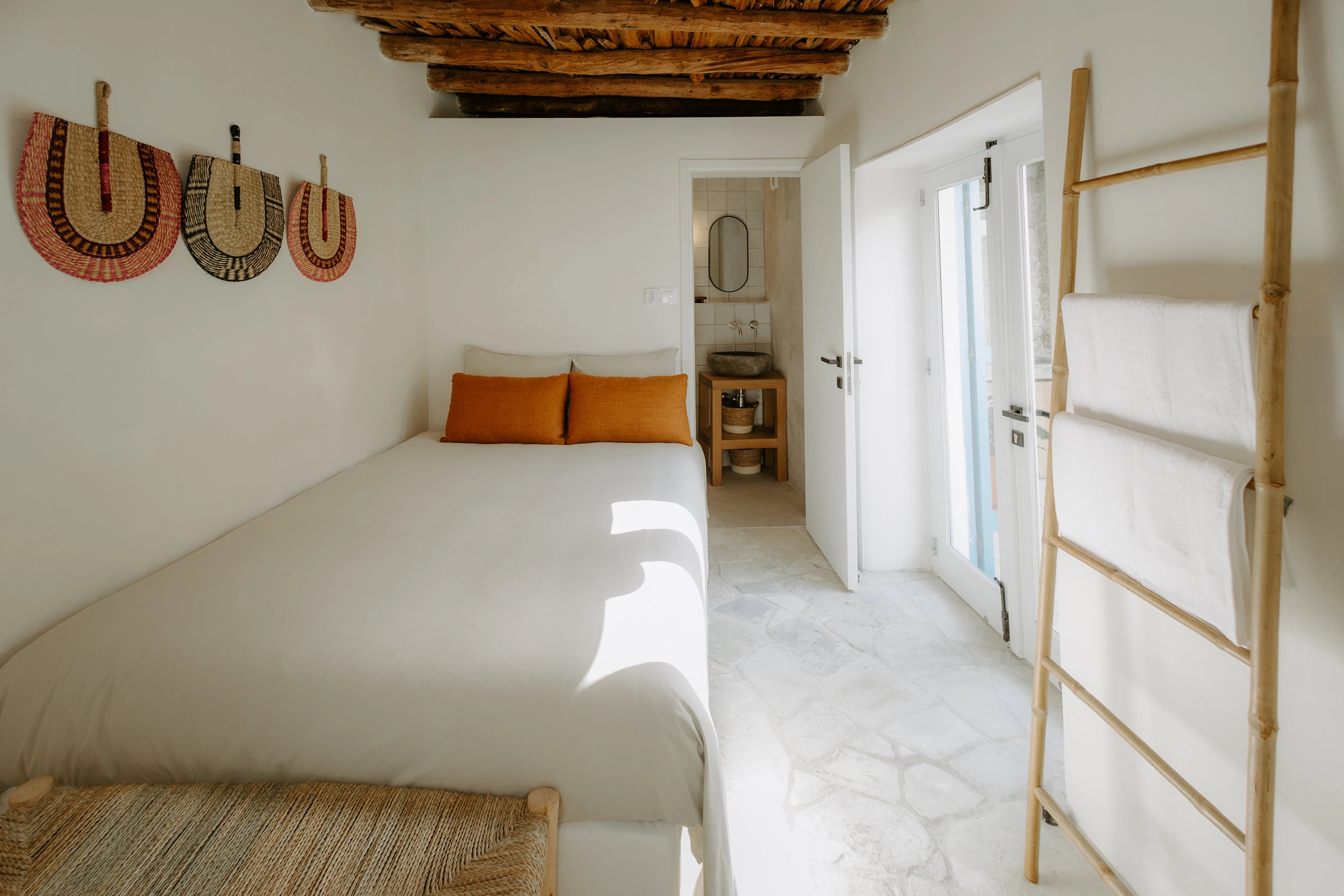 A bright bedroom with exposed timber ceiling beams, a white bed and woven wall decorations.