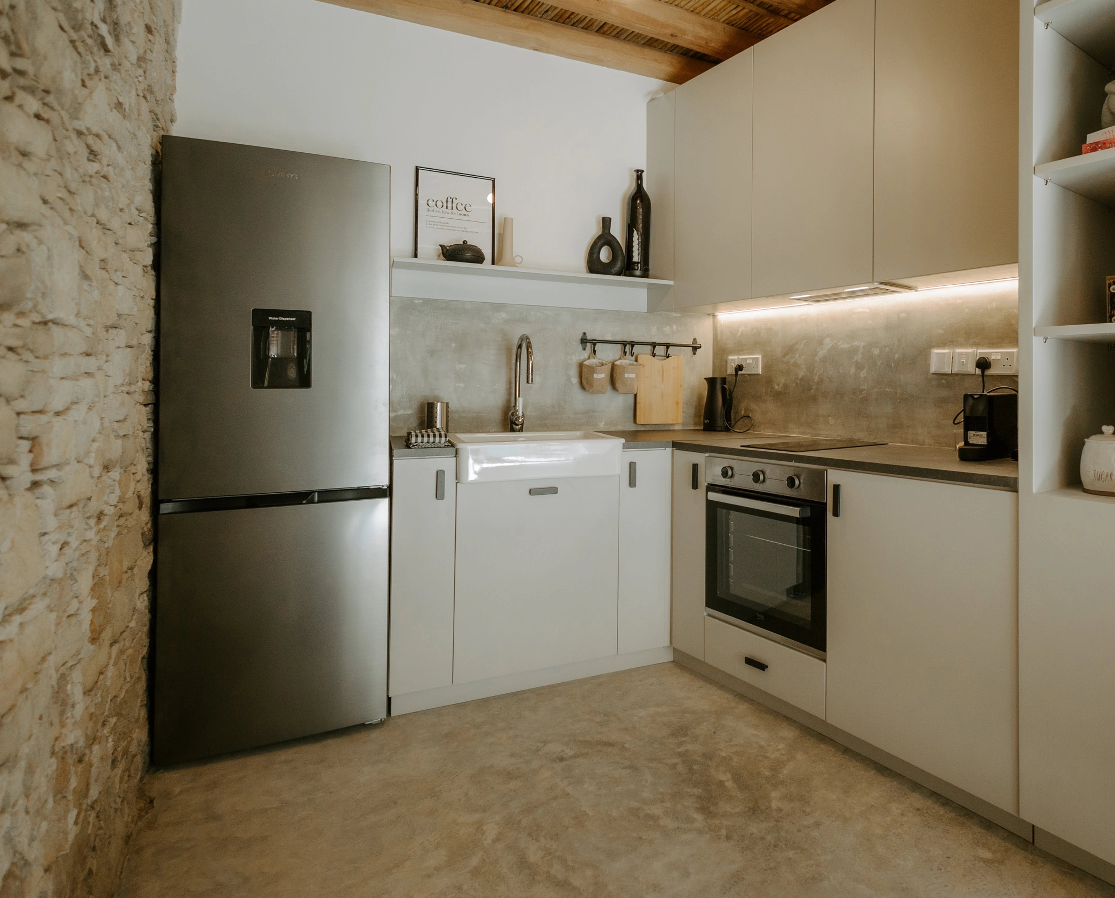 A compact pale kitchen with exposed stone walls, timber ceiling beams and a stainless-steel fridge.